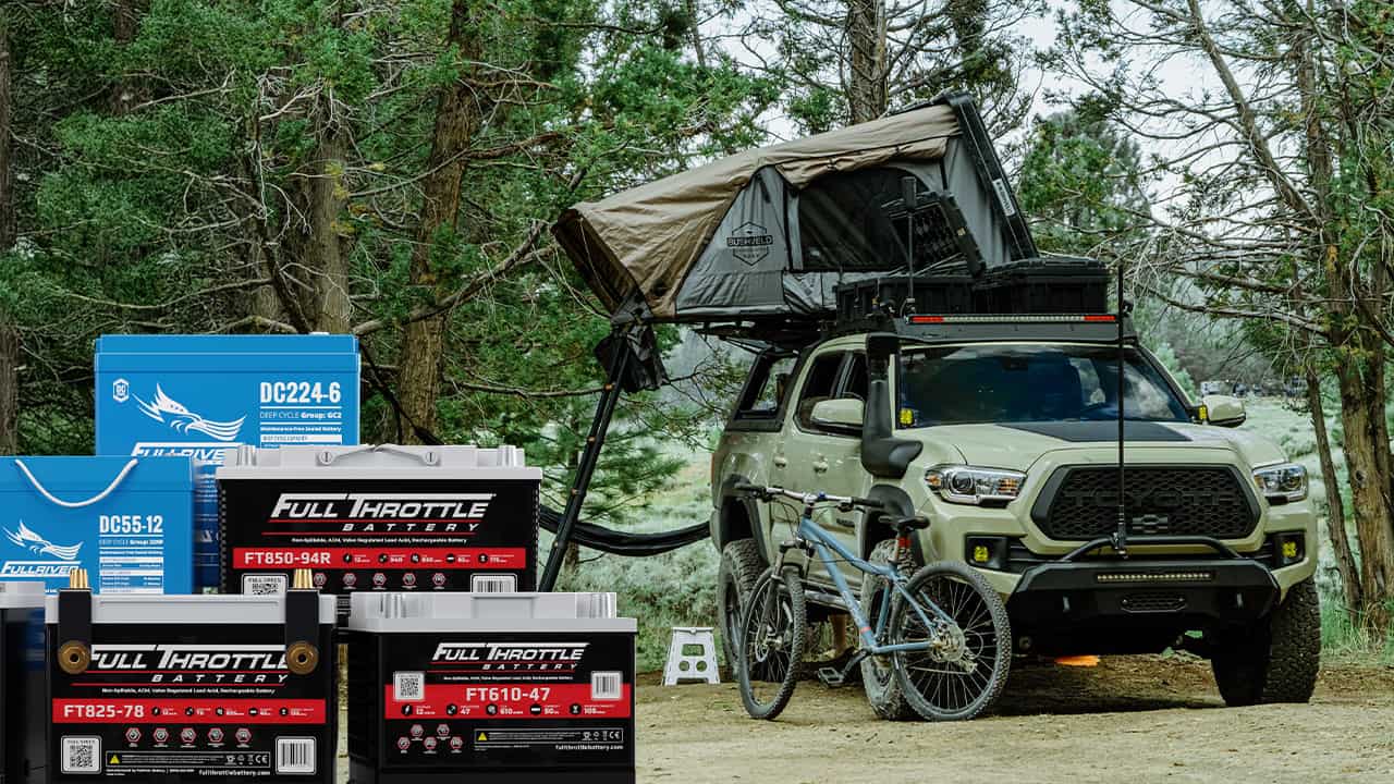 Off-road vehicle with rooftop tent in wooded area, surrounded by trees. Two bicycles are nearby. Several battery packages are displayed in the foreground.