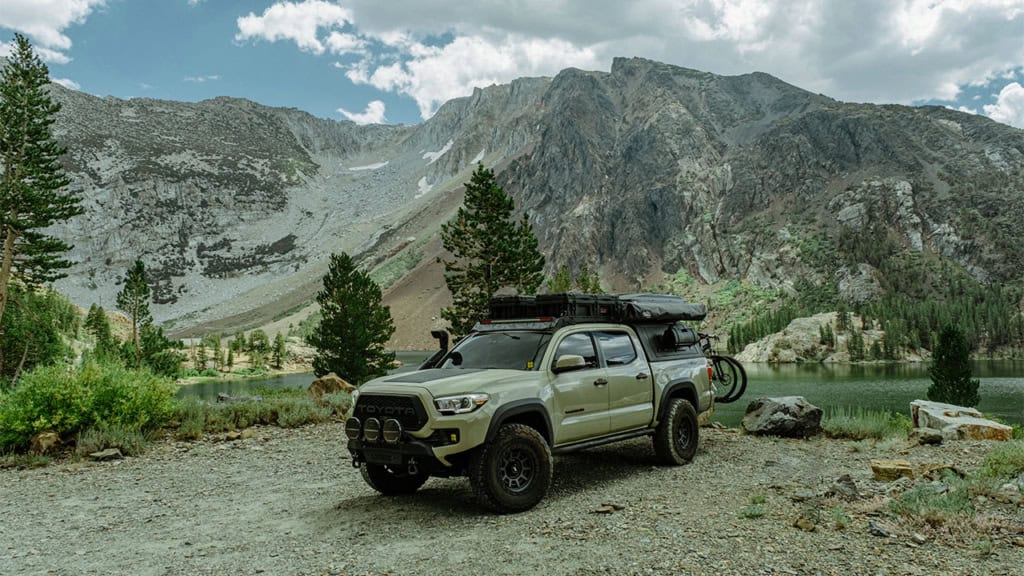 A green truck with off-road modifications is parked near a mountain lake, surrounded by trees and mountains under a cloudy sky. A bicycle is attached to the truck's rear.