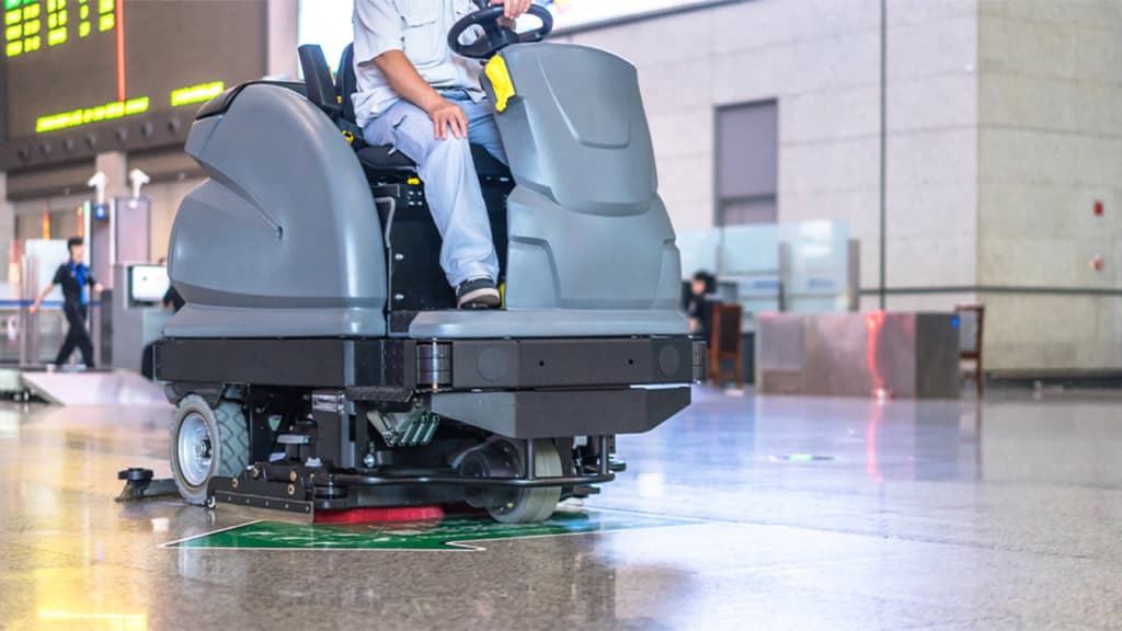 Person operating a large floor cleaning machine in a spacious indoor area with polished floors.
