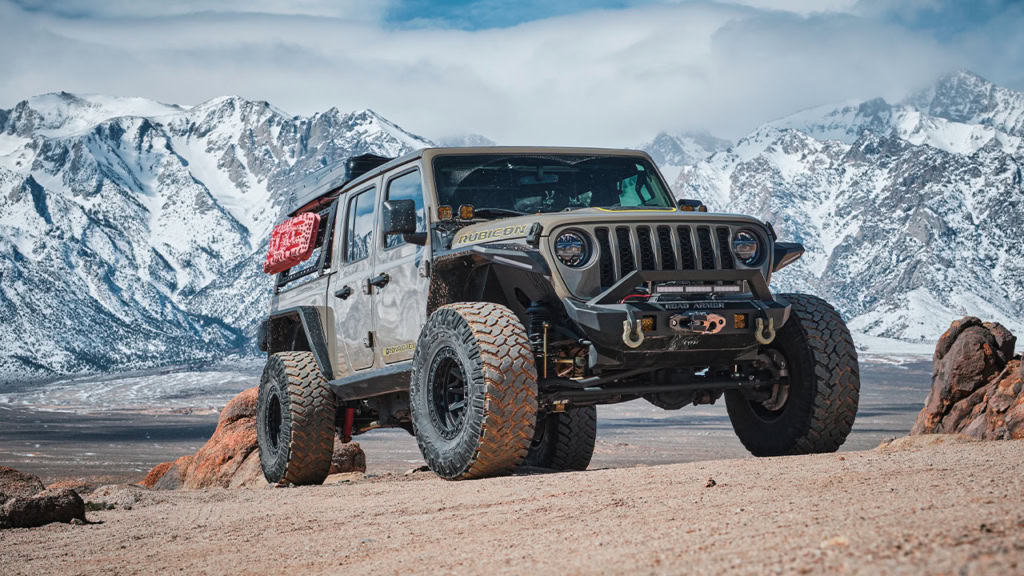 A rugged off-road vehicle on rocky terrain with snow-capped mountains in the background under a cloudy sky.