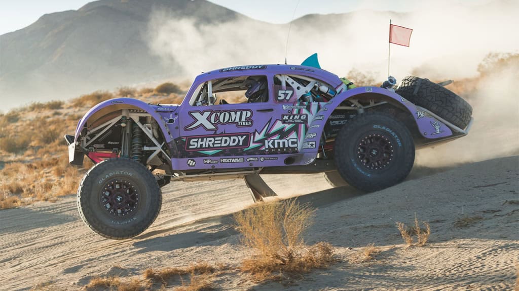 A purple off-road race vehicle with "XCOMP" and "SHREDDY" logos speeds through a desert terrain, kicking up dust. It has large tires and an exterior roll cage. A red flag is visible at the rear.