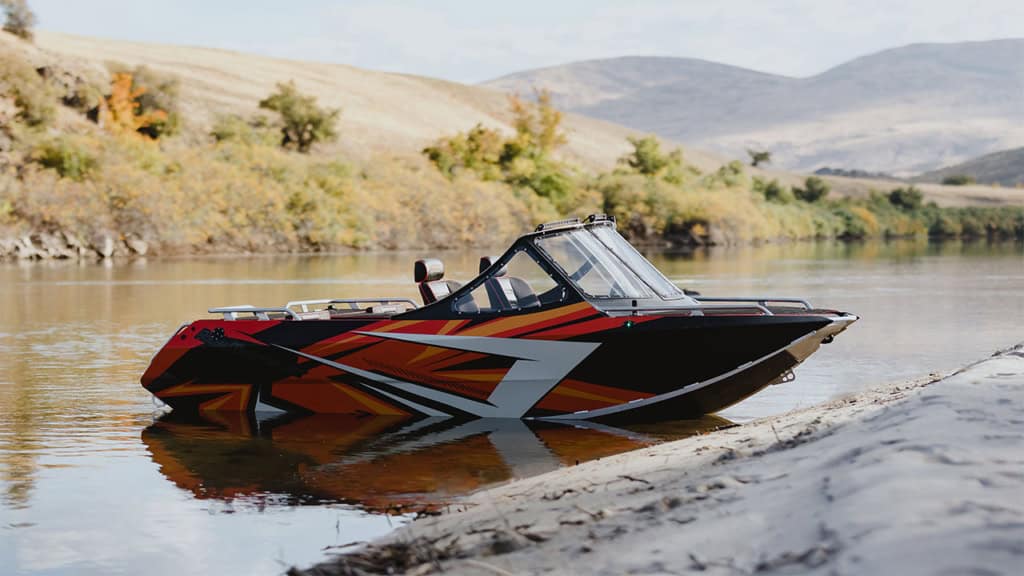 A black and red speedboat with bold geometric designs is parked on the edge of a calm river, surrounded by hills and autumn foliage.